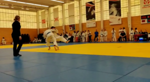A judo competition match with a competitor executing a throw on their opponent in a dojo setting, surrounded by spectators and coaches.