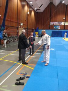 A judo practitioner in a white gi shakes hands with two officials in a gymnasium, with a judo mat visible in the background.